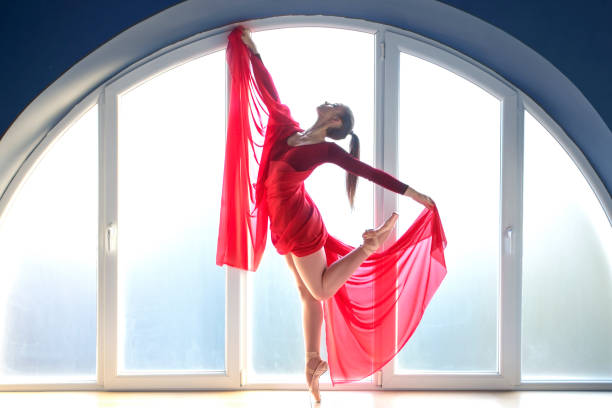 belle jeune ballerine concentrée dans une élégante longue robe de ballet rouge en pose papillon se tient devant la grande fenêtre de forme ronde du studio de ballet tandis que la lumière naturelle du jour illumine l’intérieur avec la lumière ambia - round toe shoes photos et images de collection