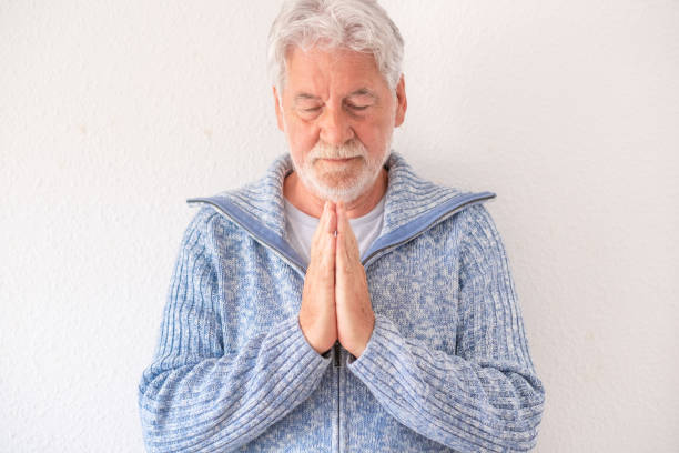 istock Bearded senior man in casual clothes with folded hands praying standing against white background. Elderly man in blue sweater in spiritual prayer 1396115612