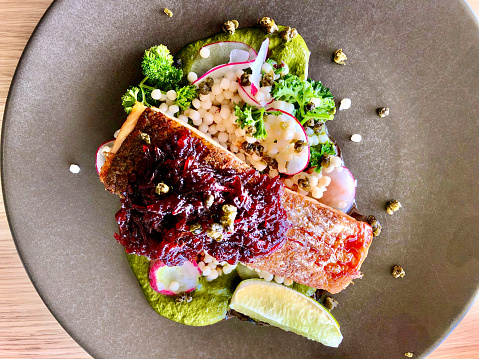 Horizontal high angle closeup photo of a slice of Tasmanian pan-fried Salmon with Pearl couscous, sliced radishes, green Basil pesto, pickled beetroot relish, parsley, capers and a lemon wedge on a grey ceramic plate on a table in a fine dining restaurant.