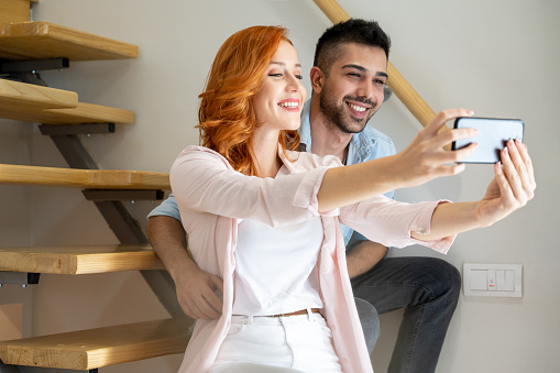 Beautiful diverse young couple sitting on stars at home and taking a selfie photo on smartphone. Red haired woman holding smartphone to take self portrait and embracing with her boyfriend