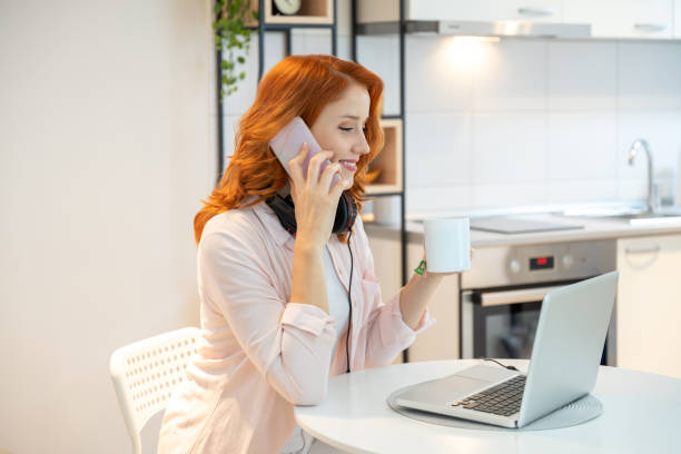 portrait d’une femme rousse souriante buvant du thé, parlant sur smartphone et utilisant un ordinateur portable dans le salon. ginger girl assise sur un bureau et ayant un appel vidéo sur ordinateur portable à la maison. - women on the phone headset service photos et images de collection