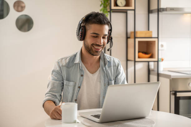 portrait d’un homme souriant portant des écouteurs avec microphone utilisant un ordinateur portable à la maison. - desk on the phone sitting table photos et images de collection