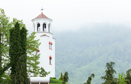 Place of power. White pretty tower with cross in the mountain, foggy green forest on the background in spring (summer) time, peaceful, beautiful landscape, high quality photo. Postcard. Horizontal plane.