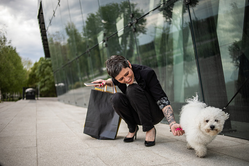 A mature woman walks past the mall, carries shopping bags and cuddles her dog