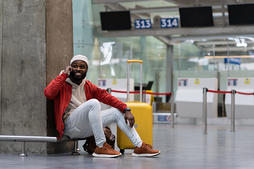Cheerful black male sitting on floor relaxing near wall airport with before announcement for flight check-in, talking on smartphone, waiting friend for together journey. Pleasure of traveling concept.