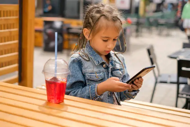 Photo of A child with Afro-pigtails in a denim jacket is playing games on a smartphone. The kid is sitting at a table in a patio in a cafe and looks intently at the phone. A girl drinks lemonade. Summer Day