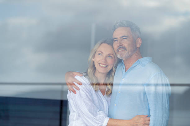 Mature couple looking at the view in their home. They look happy and contented. Mature couple looking at the view in their home. They look happy and contented. They are embracing looking through a window. wealthy house stock pictures, royalty-free photos & images