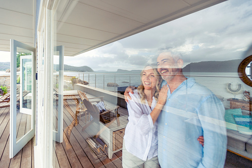 Mature couple looking at the view in their waterfront home. They look happy and contented. They are embracing looking through a window. The ocean can be seen in the reflection