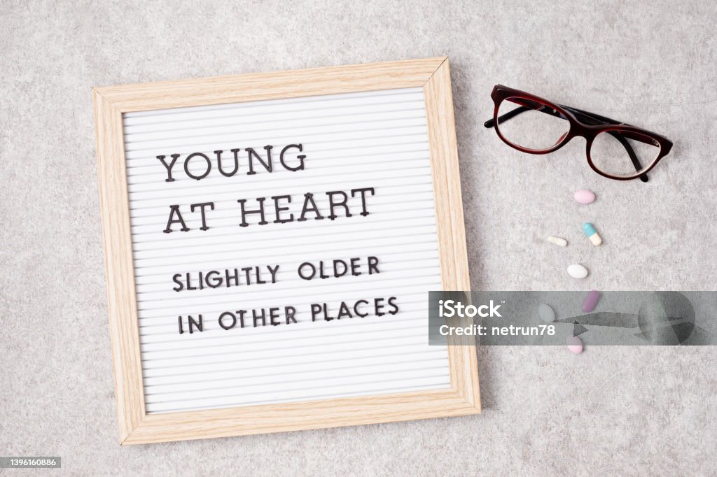 Letter board with text young at heart, slightly older in other places Letter board with text young at heart, slightly older in other places. Eye glasses and medicines on background. Ageing concept Healthcare And Medicine Stock Photo