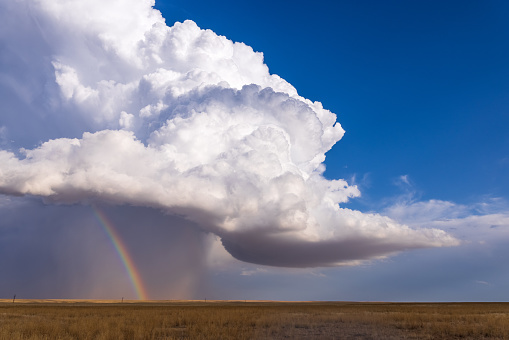 Big puffy cumulus cloud with an embedded thunderstorm in the distance on the eastern plains of Colorado. A brilliant, bright rainbow magically appears from the rain shaft