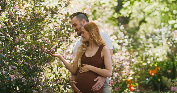 A pregnant young woman walking in nature with her husband, sharing a romantic moment. Happy couple enjoying a relaxing maternity leave outdoors