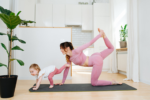 Adored mother doing yoga stance next to her baby girl on a mat in a spacious kitchen. Her cute daughter plays copycat. Wearing pink sportswear. Standing on opposite limbs.