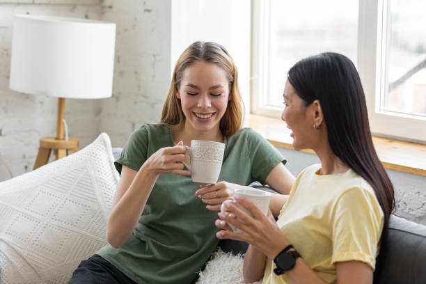 istock Two millennial girl friends at home on the sofa talking, drinking coffee, discussing. Diverse friends spending fun time together and laughing, chat and talk. Asian and Caucasian model. 1396183836
