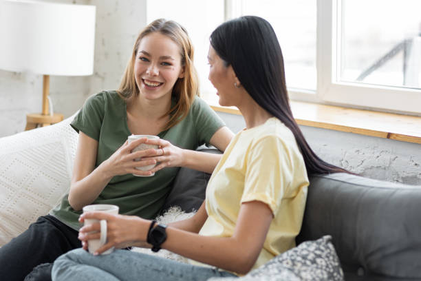 istock Two millennial girl friends at home on the sofa talking, drinking coffee, discussing. Diverse friends spending fun time together and laughing, chat and talk. Asian and Caucasian model. 1396183872