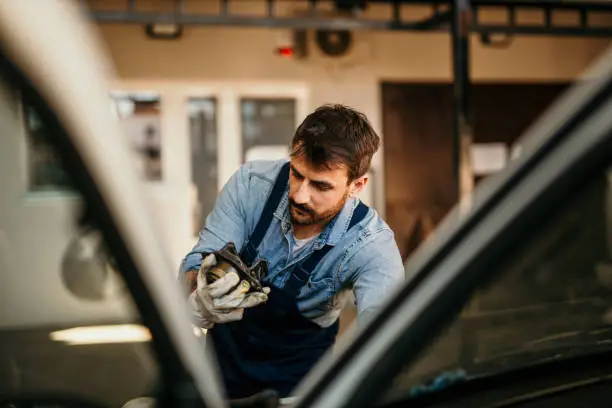 Spontaneous image of a handsome young adult, bearded, mechanic, working in the auto repair shop. He's fixing the car with focus, wearing a working outfit with a gloves. Auto car repair service center.