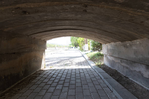 Concrete underpass extends into distance, featuring textured walls with horizontal ridges. Warm overhead lighting illuminates passageway, reflecting off wet asphalt floor. Educating local residents about the significance of flood level indicators on tunnel walls can help prevent accidents during storms