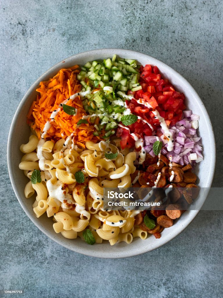 Image of bowl of Gomiti (elbow) tubular pasta, diced cucumber, grated carrot, sausage slices, chopped red onion and tomato drizzled with mayonnaise, topped with toasted seeds and nuts, elevated view Stock photo showing elevated view of a dish filled with Gomiti ridged pasta, diced cucumber, grated carrot, sausage slices, chopped red onion and tomato. Uncooked Pasta Stock Photo