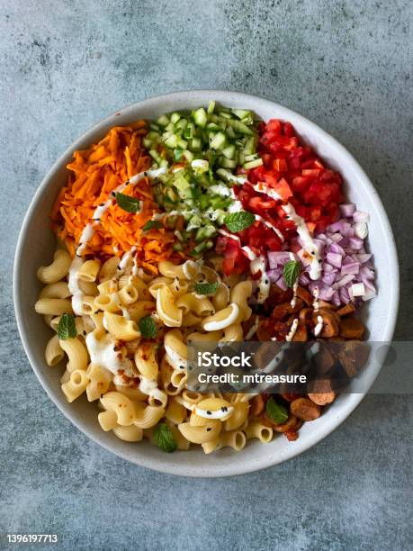 Image Of Bowl Of Gomiti Tubular Pasta Diced Cucumber Grated Carrot Sausage Slices Chopped Red Onion And Tomato Drizzled With Mayonnaise Topped With Toasted Seeds And Nuts Elevated View Stock Photo - Download Image Now