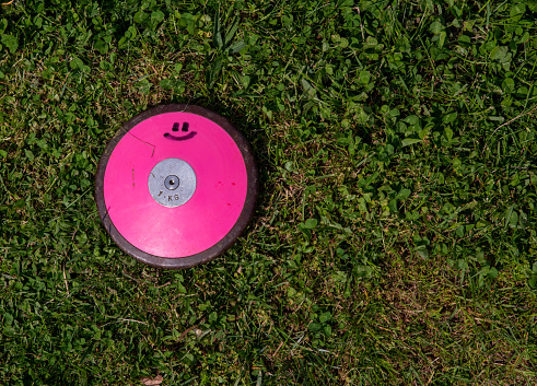 A 1 kg round pink track and field discus with a smiley face lays off center in the grass. Shot in natural sunlight with no people and copy space.