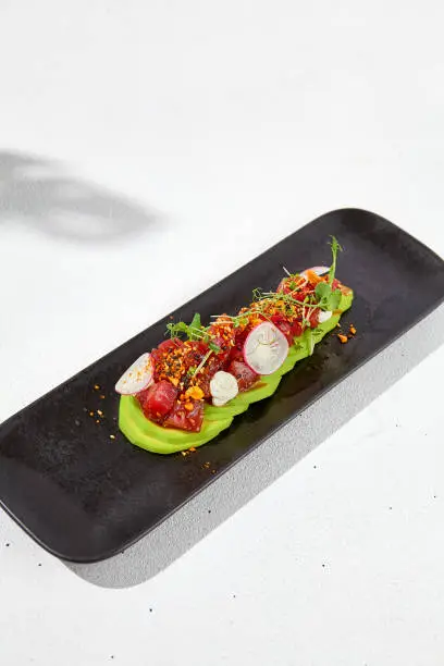 Modern food - tuna tartare on sliced avocado in black plate. Avocado starter with tuna and radish in asian style. Contemporary asian dish. Healthy food with avo and marinated fish on white background