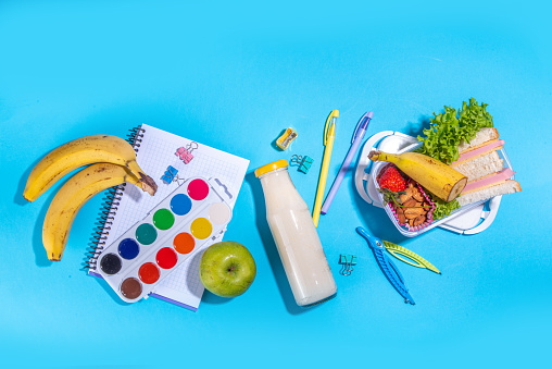 Healthy school lunch box: sandwich, vegetables, fruit, nuts and yogurt with school kids supplies, accessories and backpack on high-colored blue background flatlay copy space