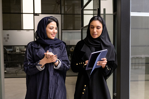 Front view of young Middle Eastern women in traditional abayas and headscarves walking together and smiling as they look at content on portable device.