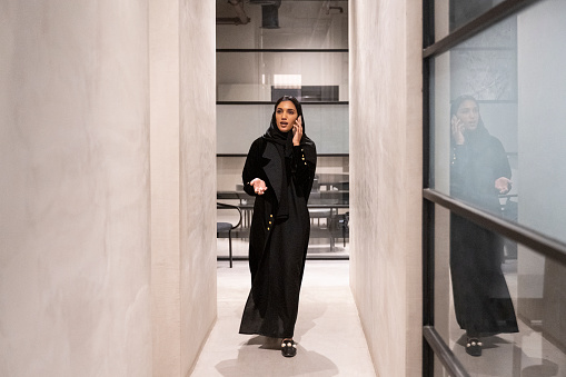 Full length front view of Riyadh professional in traditional black abaya and hijab communicating with caller while approaching camera in office hallway.