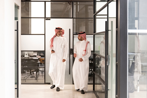Full length view of bearded men in 20s and 40s wearing traditional Saudi attire and conversing face to face as they move through work environment.