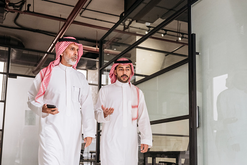 Low angle view of professionals in 20s and 40s wearing traditional Saudi attire and talking as they return to their offices from business meeting.