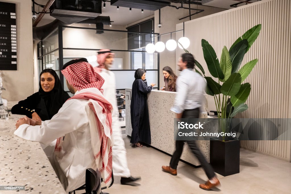 Middle Eastern professionals in Riyadh coworking office Mixed-age business people in traditional Saudi and western attire relaxing at coffee bar, walking through lobby, and talking at reception desk. Saudi Arabia Stock Photo