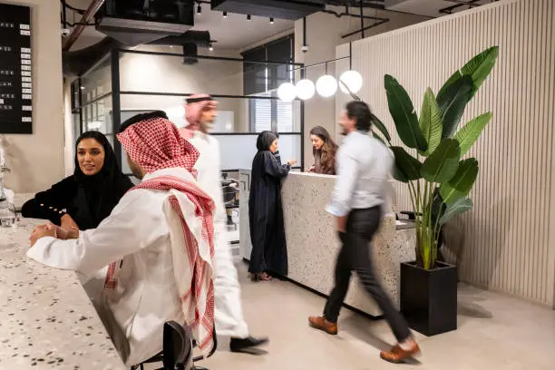 Mixed-age business people in traditional Saudi and western attire relaxing at coffee bar, walking through lobby, and talking at reception desk.