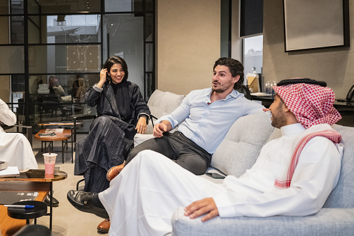 Middle Eastern men and women in traditional and western attire sitting in modern office lounge talking and laughing.