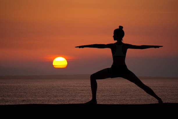 Woman silhouette in Yoga Warrior pose at sunset with quiet ocean Woman silhouette in Yoga Warrior pose at sunset with quiet ocean in background yoga stock pictures, royalty-free photos & images
