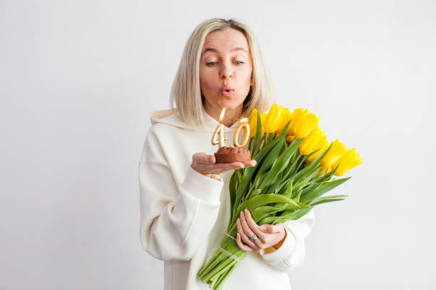 Portrait of happy middle aged woman holding birthday cake with lit candles Middle-aged woman blows out the candles on the cake. Female with bouquet of tulips on wall background woman birthday cake stock pictures, royalty-free photos & images