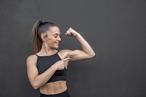 Strong sportswoman with ponytail smiling and pointing at muscle of bent arm while standing against black wall in gym
