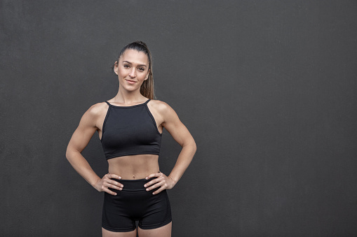 Self assured strong sportswoman in black shorts and crop top holding hands on waist and looking at camera while standing against black wall during training in gym
