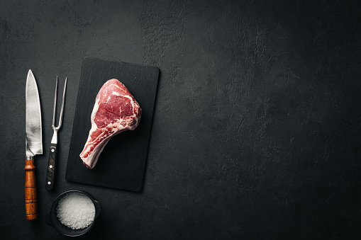 Raw tomahawk or cowboy steak on graphite cutting board over black stone background. Top view, copy space
