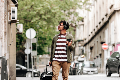 Handsome young adult man with suitcase walking on city street and listening to music with bluetooth headphones connected to his smart phone. He is happy and smiled.