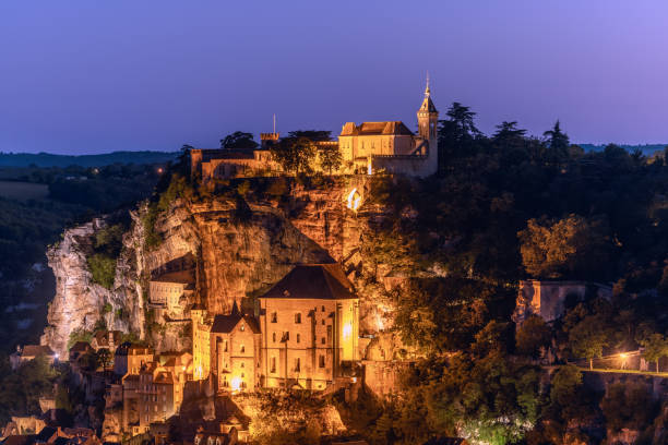 Basilica Saint-Sauveur below and Rocamadour Castle above solid rock illuminated after sunset. Lot, Occitania, Southwestern France