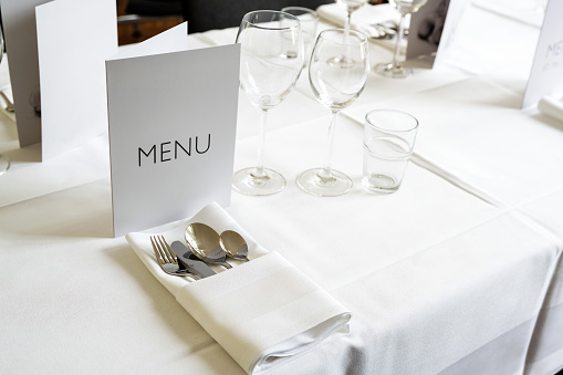 Elegant place setting with menu card, various glasses, cutlery and napkin on a table with white tablecloth for a festive dinner, copy space, selected focus, narrow depth of field