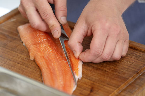 Hands of a cook remove the belly flap with bones of a fresh raw char fish with a filleting knife on a cutting board, selected focus stock photo