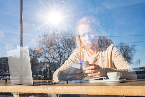 View through window of a mid adult woman looking at mobile phone in coffee bar