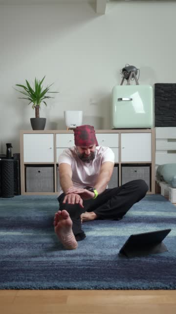 Middle-aged, bearded man doing stretches sitting on a carpet in his living room following the recommendations of an expert on his digital tablet