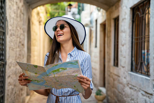 Cheerful woman with trendy look searching direction on location map while traveling abroad in summer, happy female tourist searching road to hotel during vacation