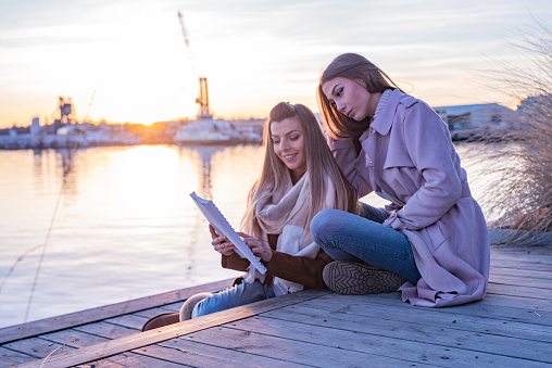 Two females friends sitting on the pier and reading a scripts