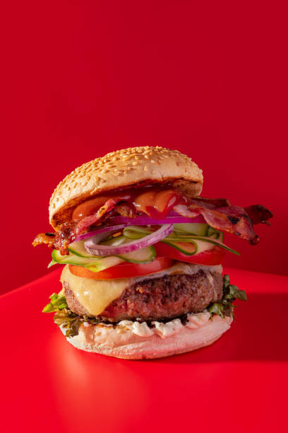 istock Bold fresh cheeseburger loaded with lettuce, cucumber, tomato and bacon on vibrant red 1396221346