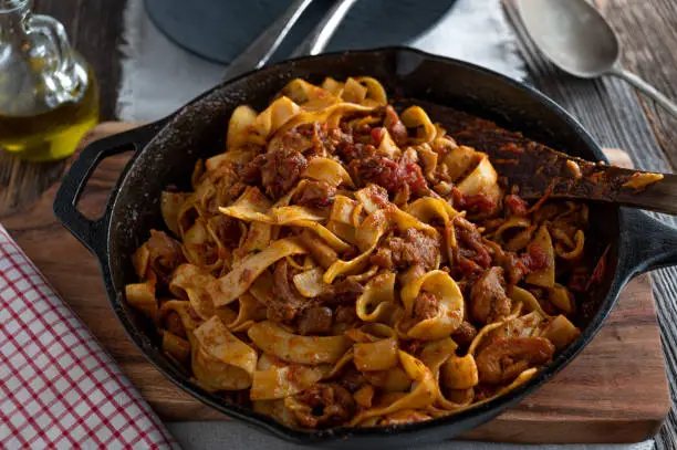 Delicious italian dish with braised chicken meat, tomato, herb sauce and tagliatelle. Served in a rustic cast iron pan or skillet on wooden table background. Ready to eat