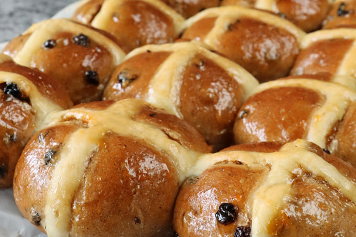 Stock photo showing close-up, elevated view of greaseproof paper covered cooling rack containing freshly baked, homemade Easter hot cross buns, home baking concept.\nThese traditional spiced sweet buns are made and sold over the Easter period, with the cross symbol on the glazed top being made from a flour and water paste, and symbolising Christianity.