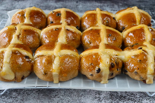 Stock photo showing close-up, elevated view of greaseproof paper covered cooling rack containing freshly baked, homemade Easter hot cross buns, home baking concept.
These traditional spiced sweet buns are made and sold over the Easter period, with the cross symbol on the glazed top being made from a flour and water paste, and symbolising Christianity. imagen en primer plano de bollos de cruz calientes de pascua recién horneados y caseros en una rejilla de enfriamiento cubierta de papel pergamino a prueba de grasa, fondo gris moteado, concepto de horneado casero, vista elevada, enfoque en primer plano