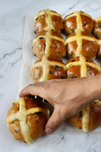 Stock photo showing elevated view of batch of freshly baked, homemade Easter hot cross buns in rows on greaseproof parchment paper, home baking concept.
These traditional spiced sweet buns are made and sold over the Easter period, with the cross symbol on the glazed top being made from a flour and water paste, and symbolising Christianity. imagen de primer plano de una persona irreconocible eligiendo un bollo con especias de filas de bollos de cruz calientes de pascua recién horneados y caseros en papel de pergamino a prueba de grasa, fondo de efecto mármol, concepto de horneado casero, vi
